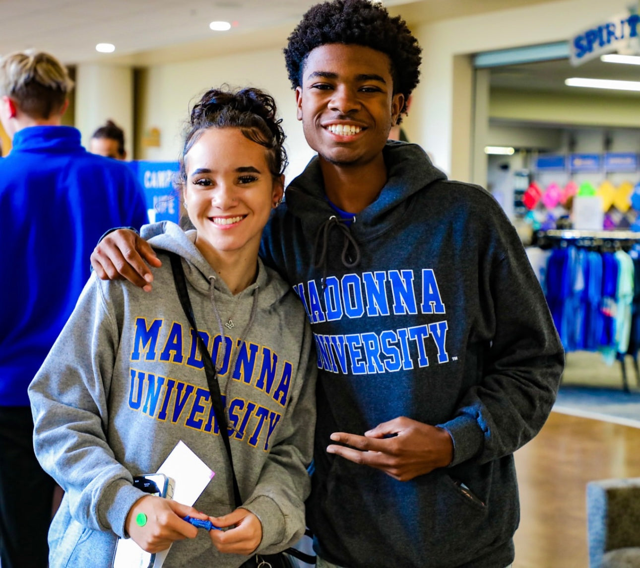 Two individuals wearing Madonna University hoodies in a casual indoor setting.