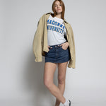 Person wearing a beige jacket, white t-shirt with text, blue shorts, and black sneakers on a plain background