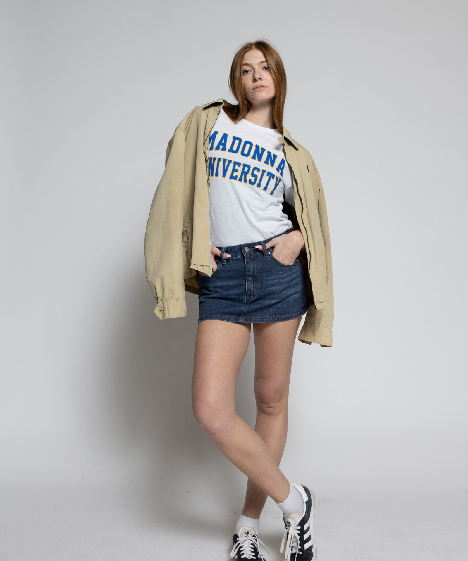 Person wearing a beige jacket, white t-shirt with text, blue shorts, and black sneakers on a plain background