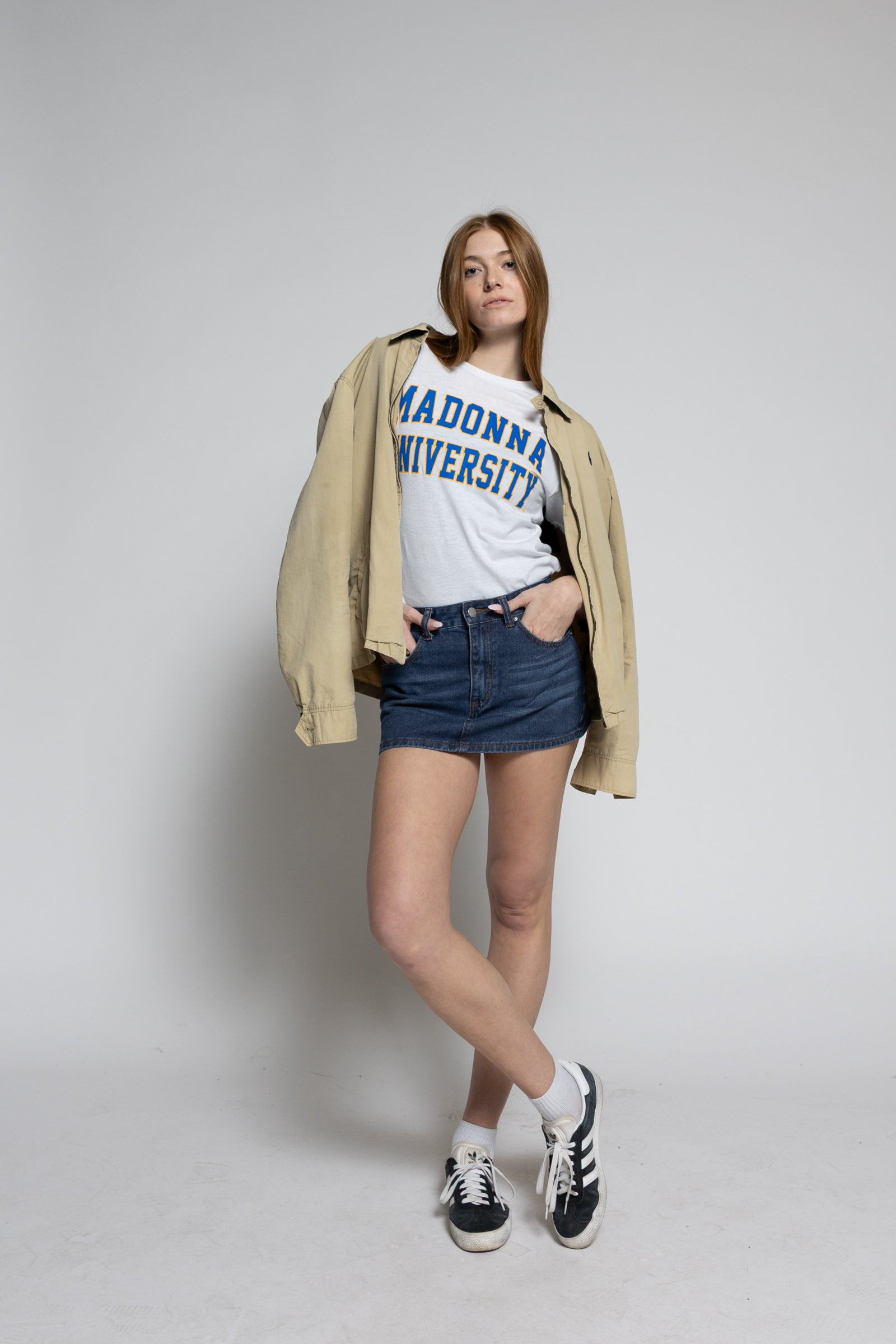 Person wearing a beige jacket, white t-shirt with text, blue shorts, and black sneakers on a plain background