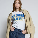 Person wearing a white t-shirt with 'MADONNA UNIVERSITY' text, denim shorts, and a beige jacket on a plain background