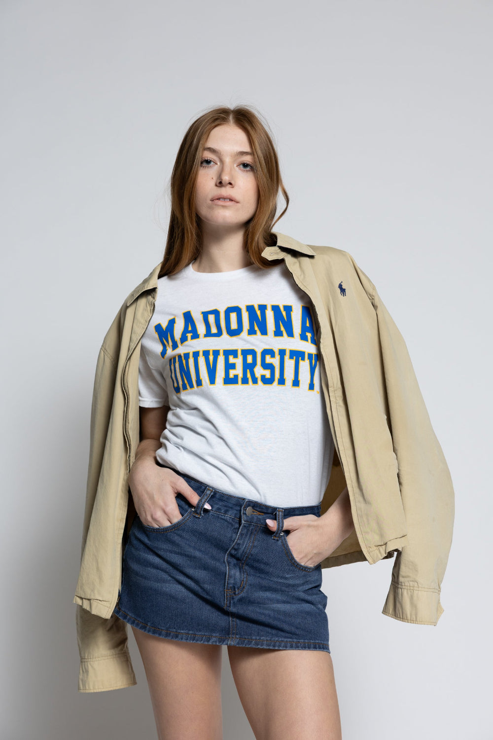 Person wearing a white t-shirt with 'MADONNA UNIVERSITY' text, denim shorts, and a beige jacket on a plain background